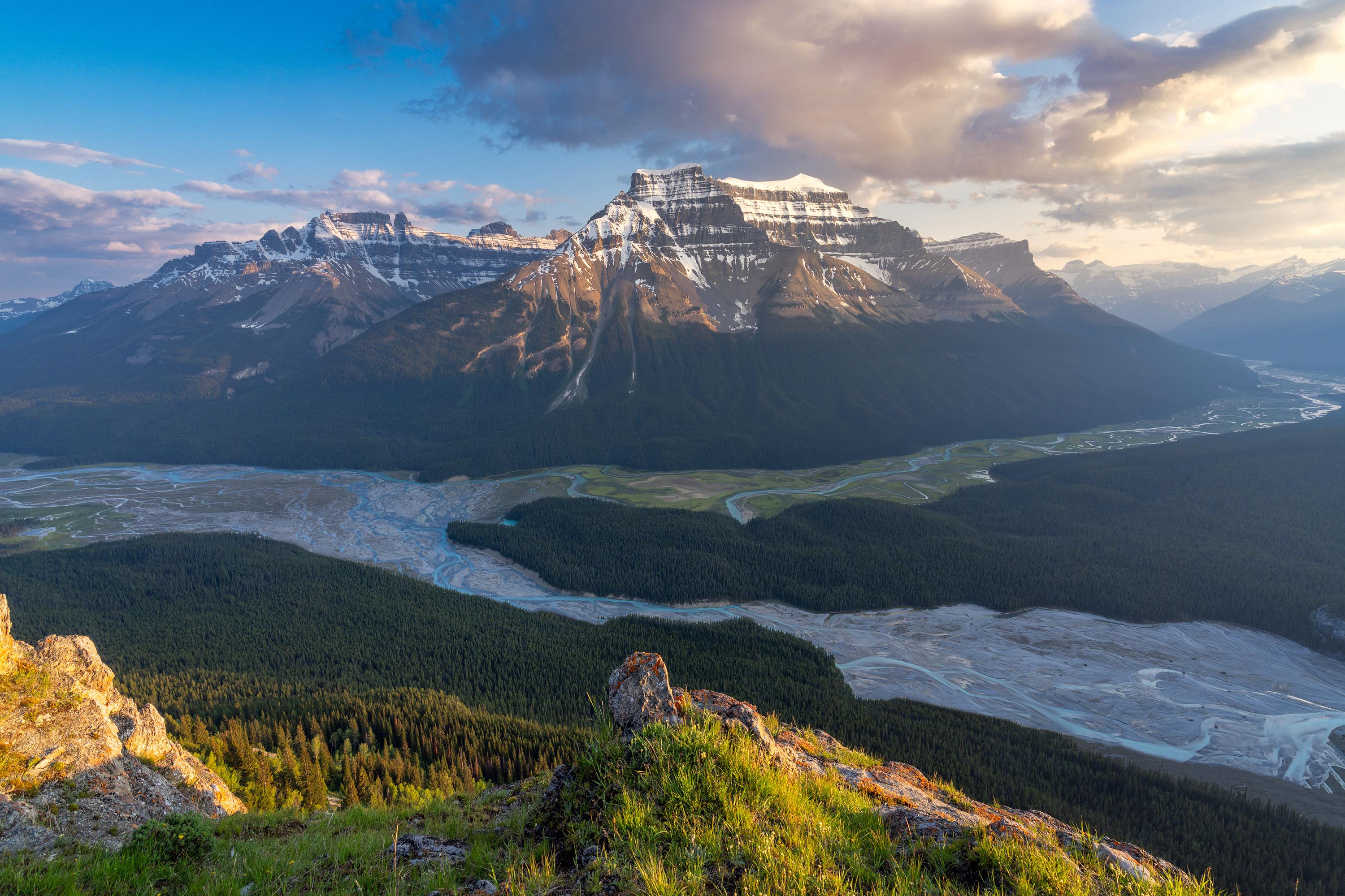 Snow-capped mountain range overlooking a forested valley and river HD Wallpaper