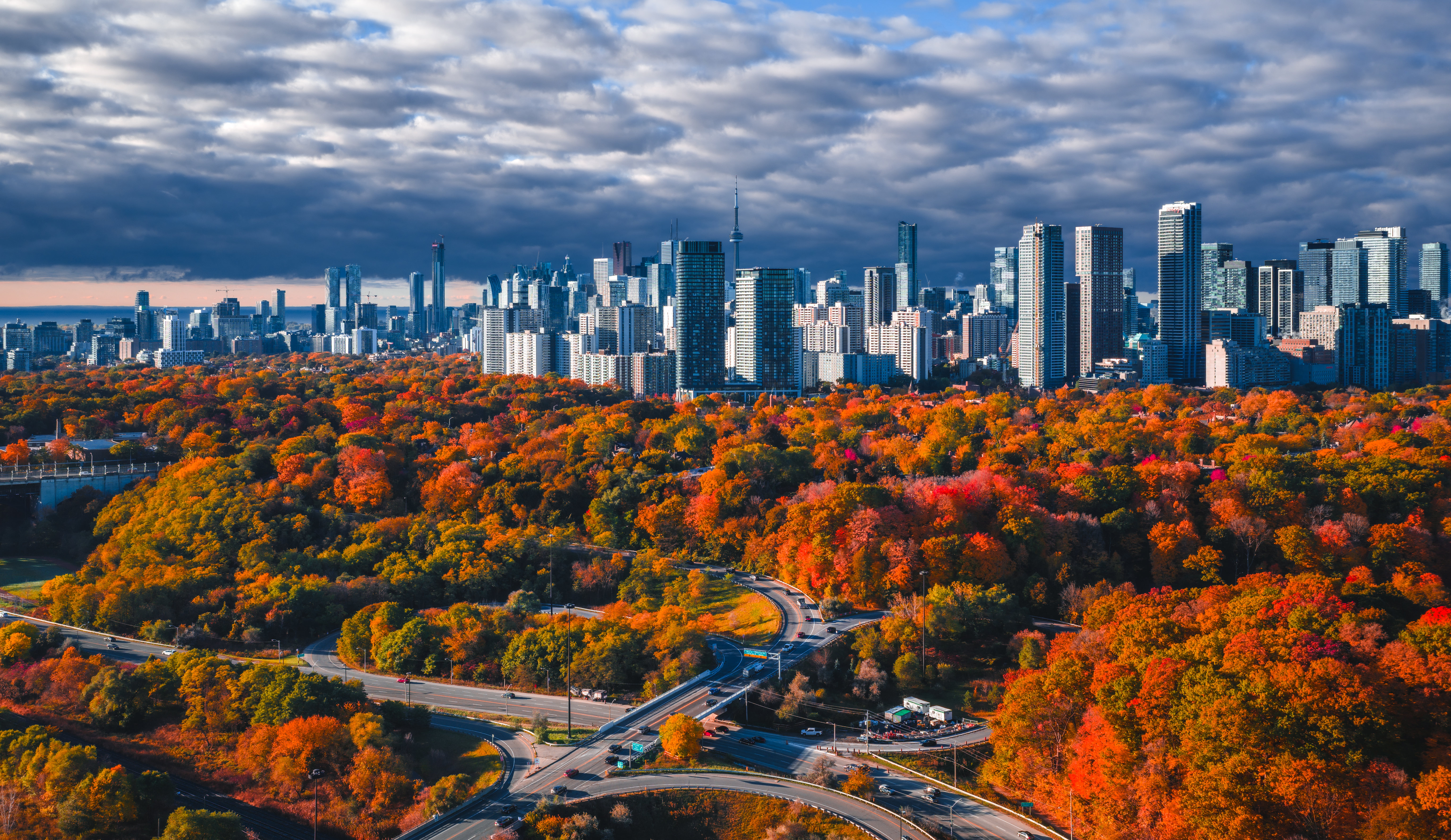 Cars on a highway surrounded by autumn trees and city skyline HD Wallpaper