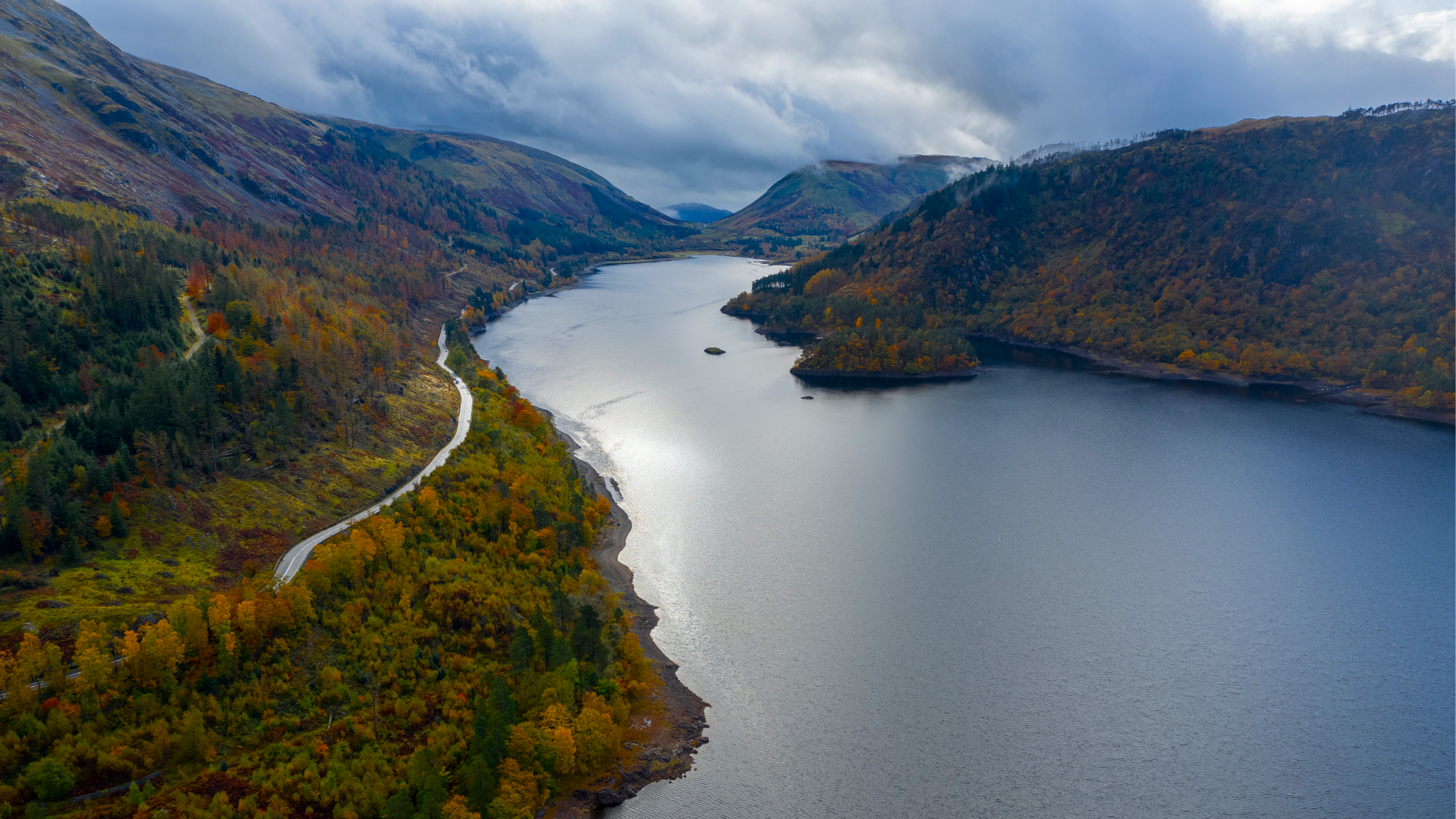 Autumn trees line a winding road next to a large lake HD Wallpaper