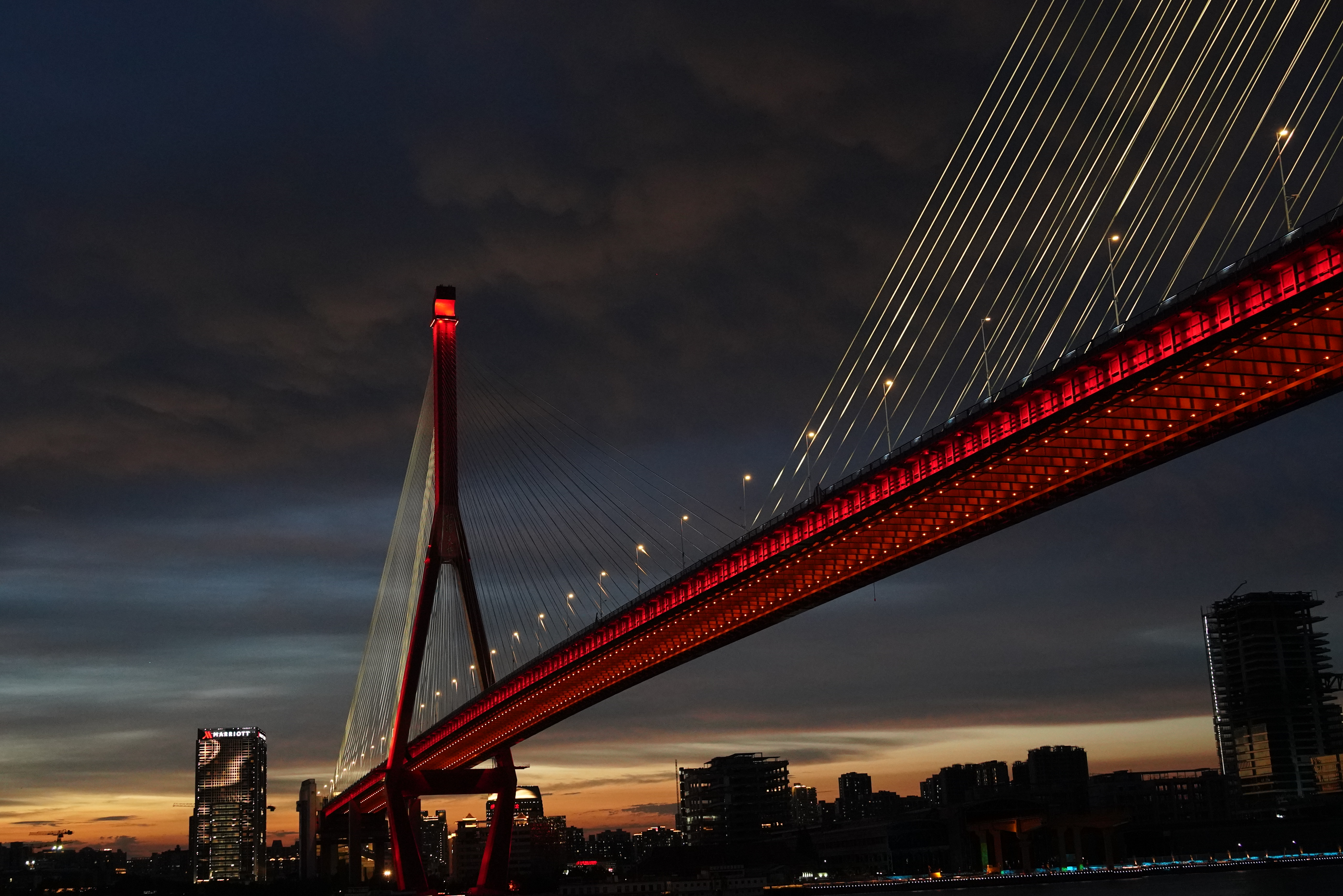 Red illuminated cable-stayed bridge at dusk over a city HD Wallpaper