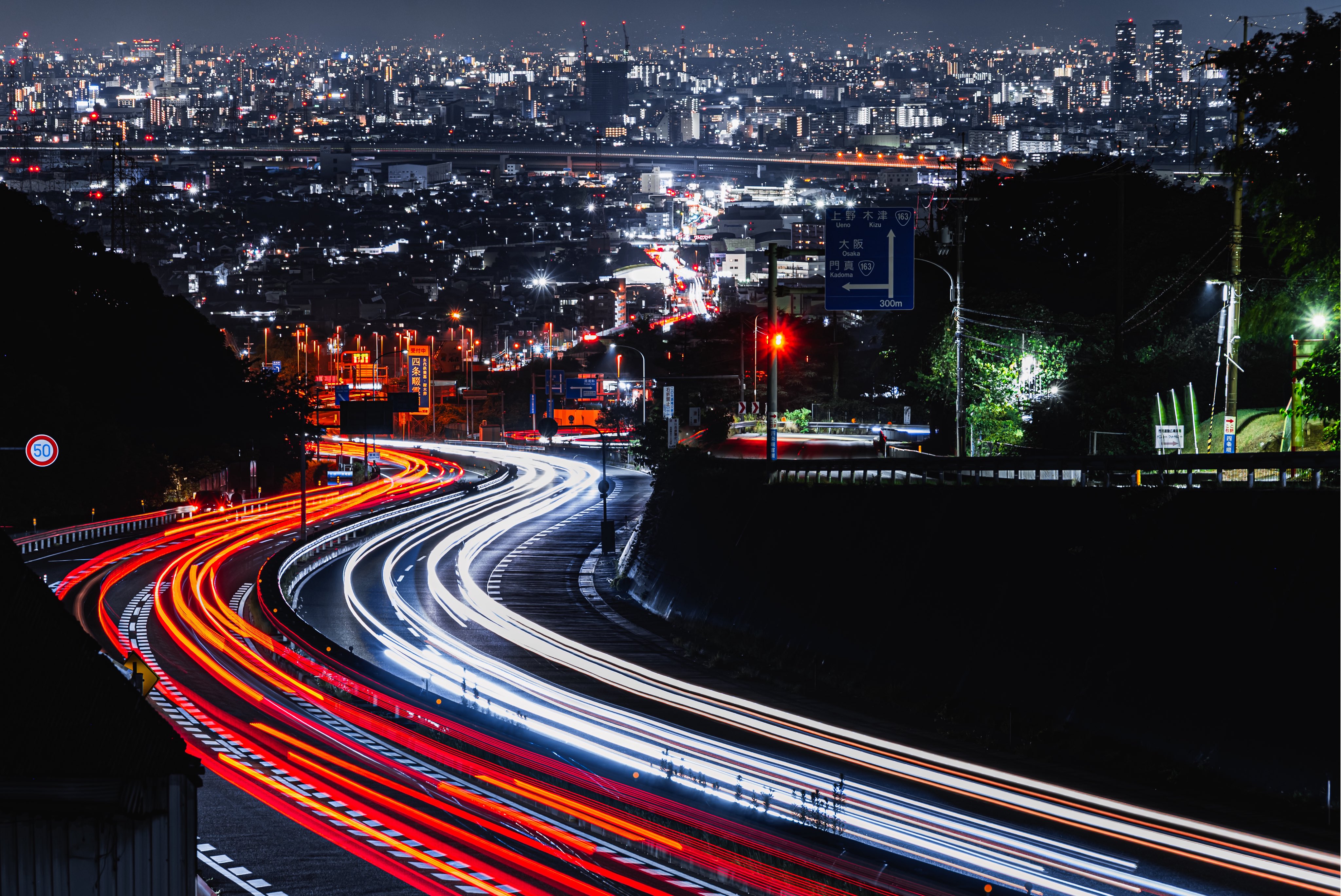 Light trails on a winding road overlooking a city at night HD Wallpaper