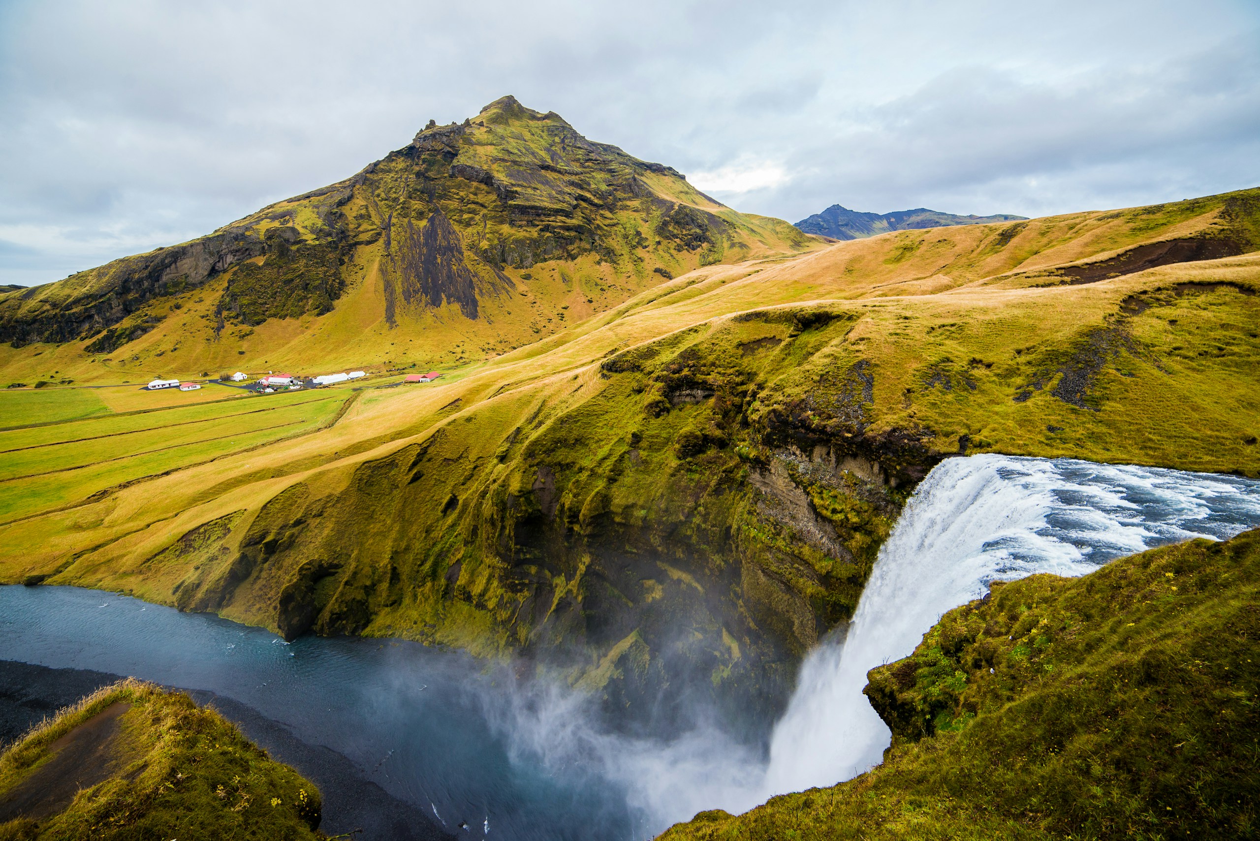 Waterfall flowing down green cliffs with mountain background HD Wallpaper