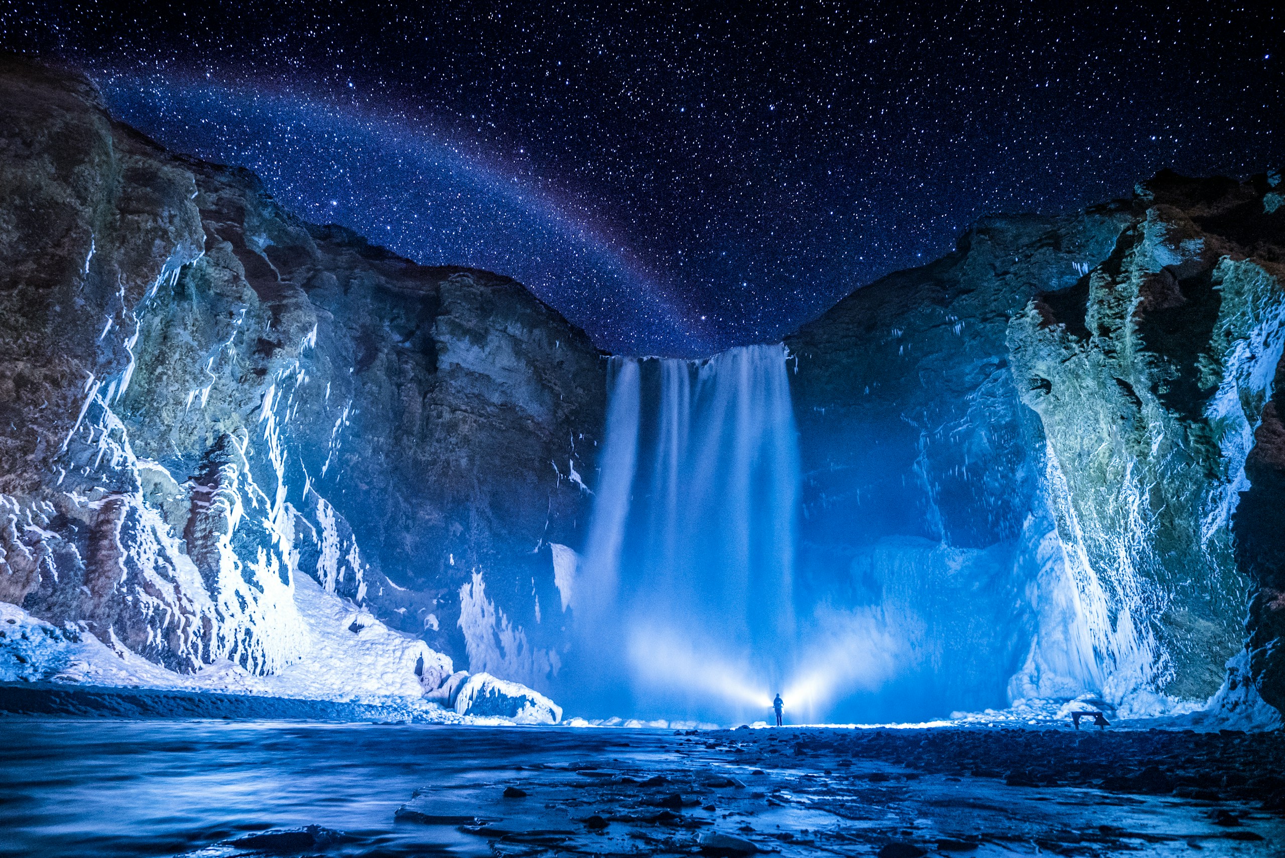 Person with flashlight at Skógafoss waterfall under stars HD Wallpaper
