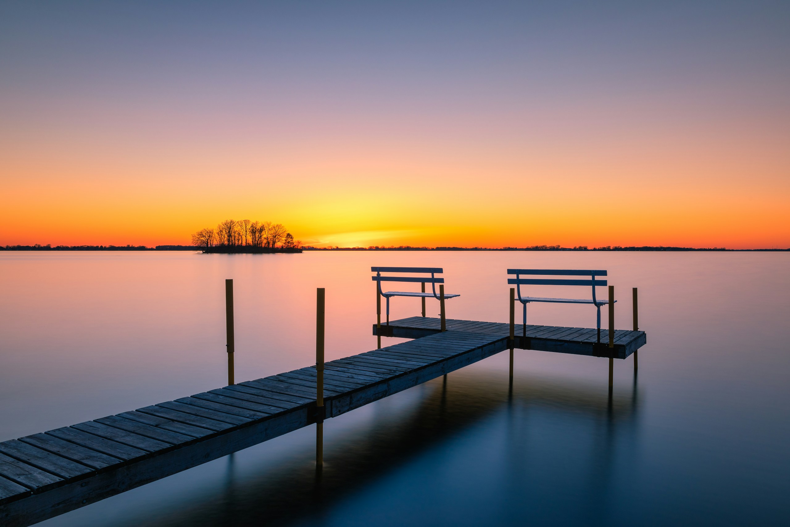 Wooden dock with benches on calm water at sunset HD Wallpaper