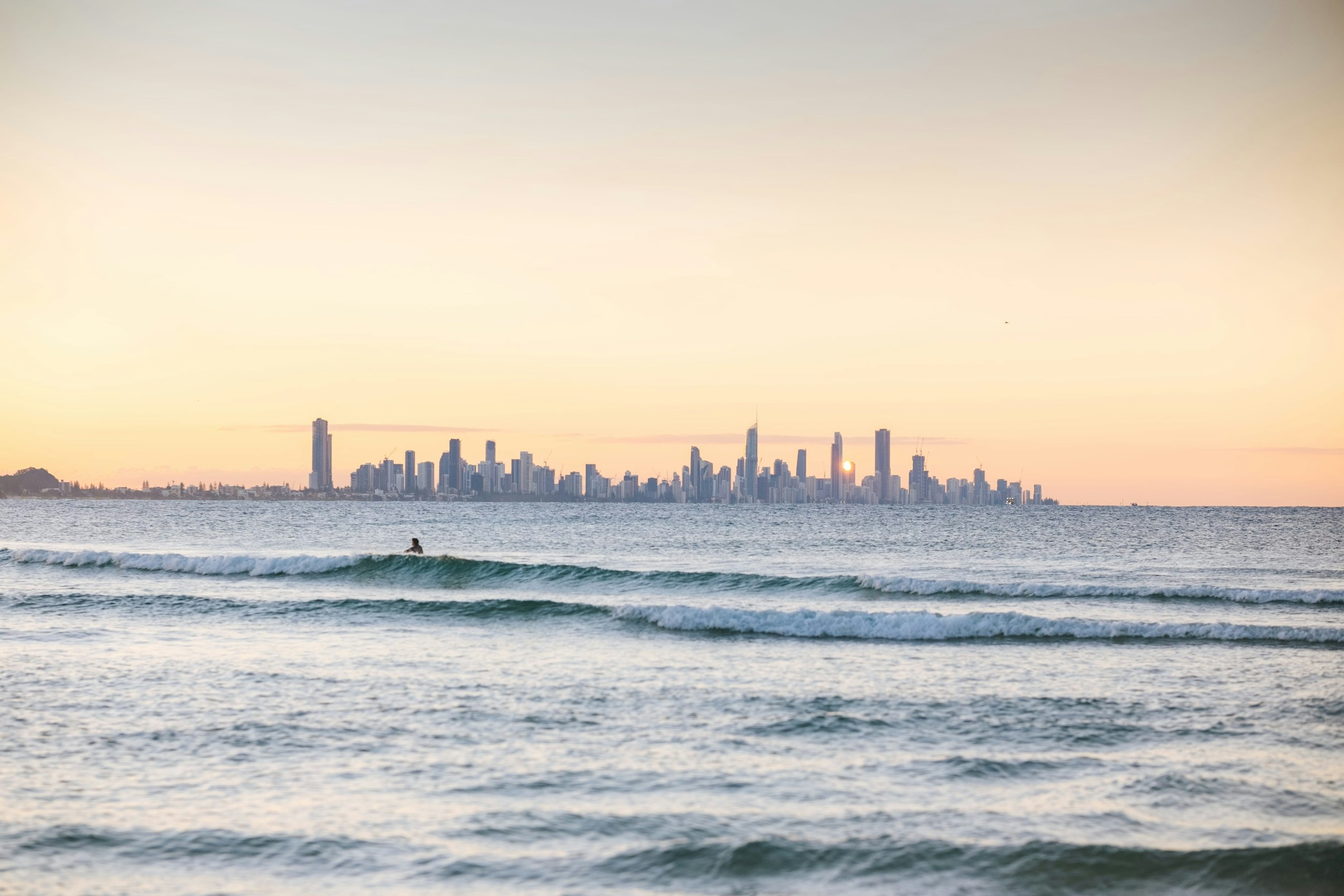 Surfer in ocean waves with city skyline at sunset HD Wallpaper