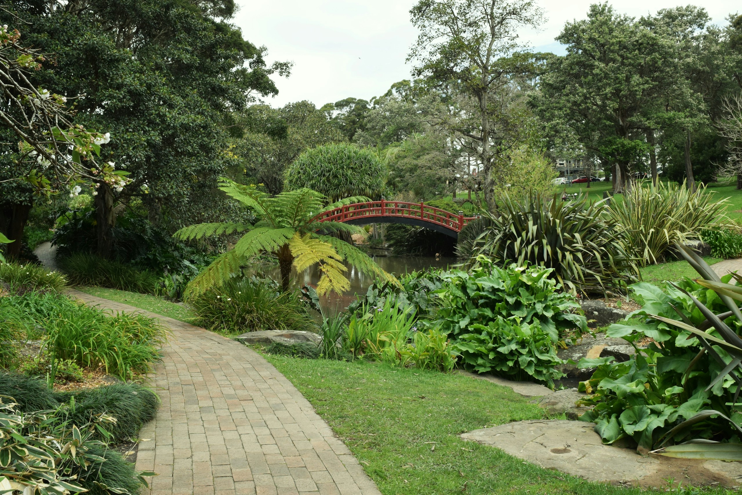 A winding brick path through a lush garden with a red bridge HD Wallpaper