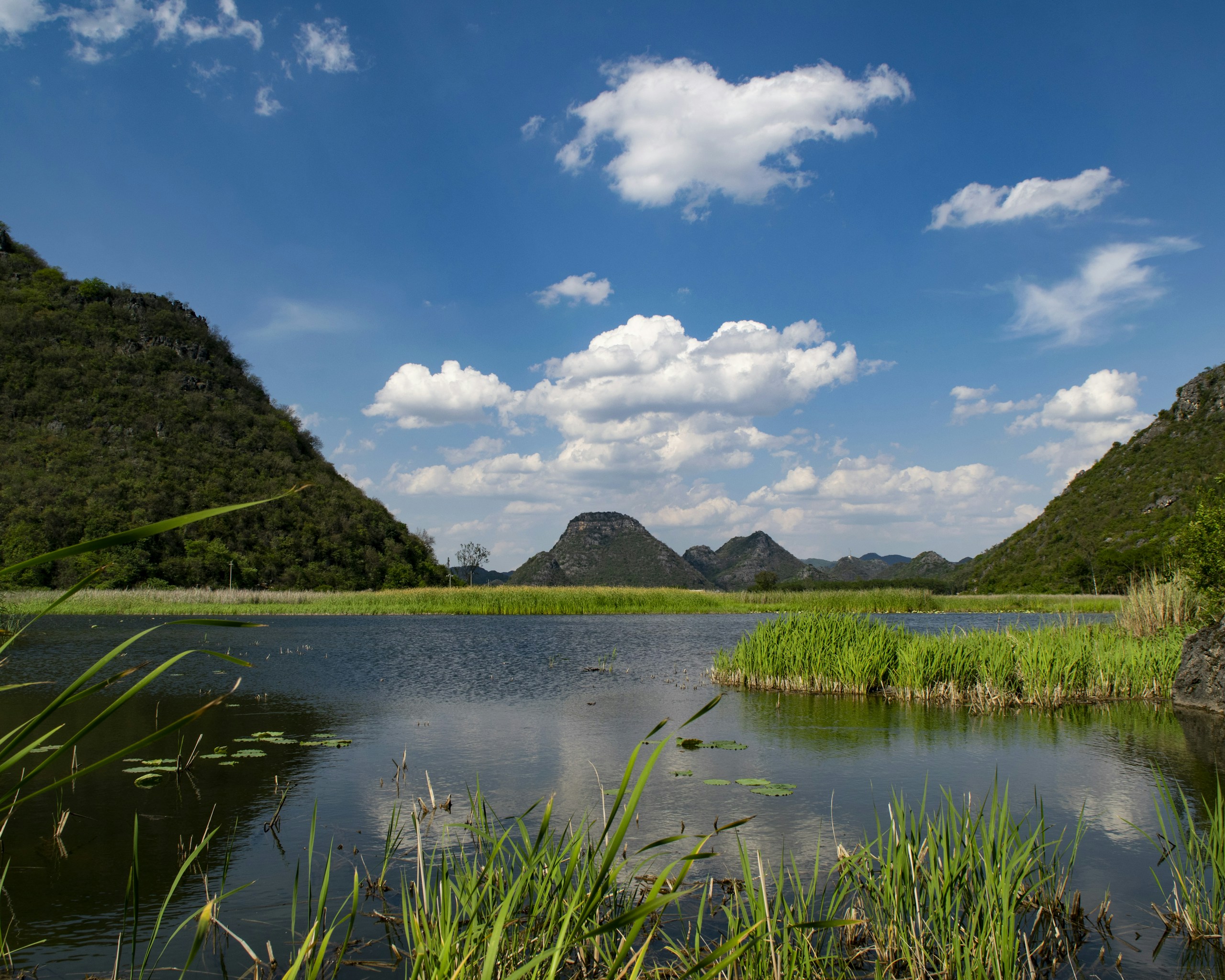 Lush green hills surround a tranquil lake under a blue sky. HD Wallpaper