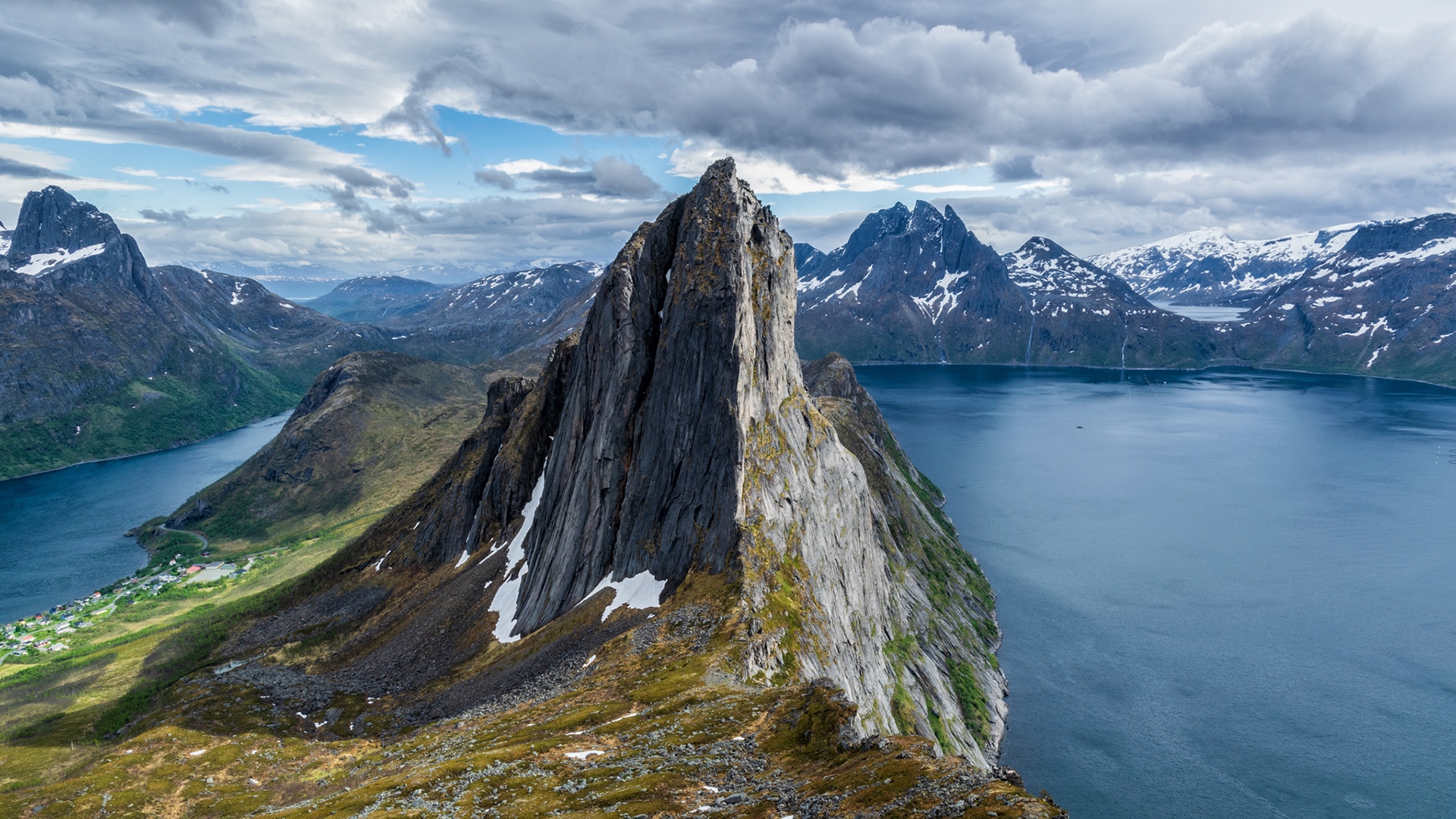 Jagged mountain peak overlooking a blue fjord and village HD Wallpaper