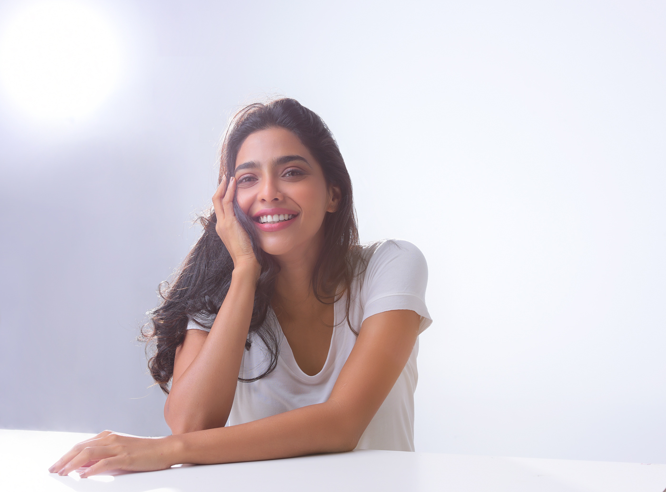 Aishwarya Lekshmi smiling, sitting at a table HD Wallpaper