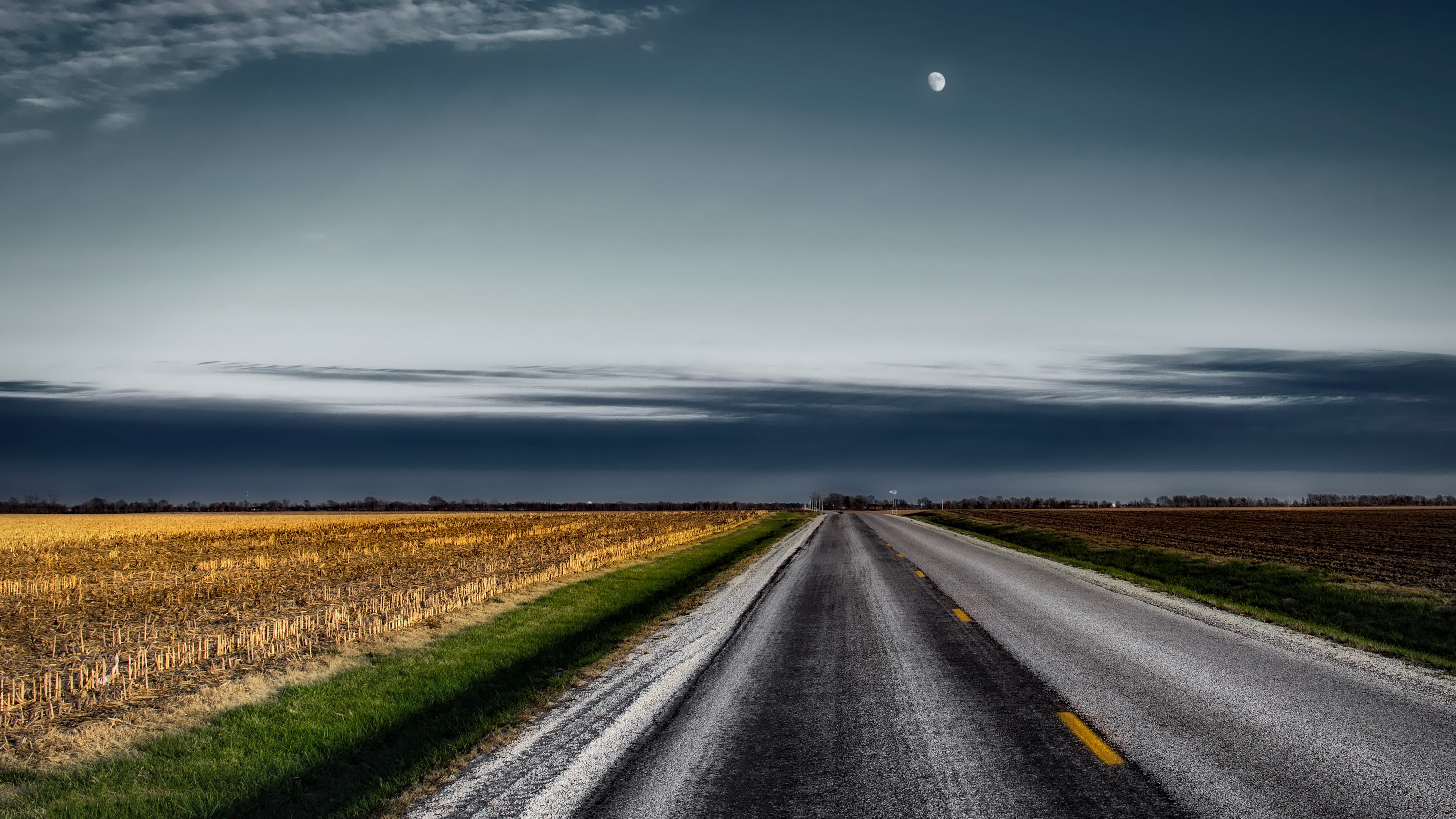 Road through a harvested field under a cloudy sky with the moon HD Wallpaper