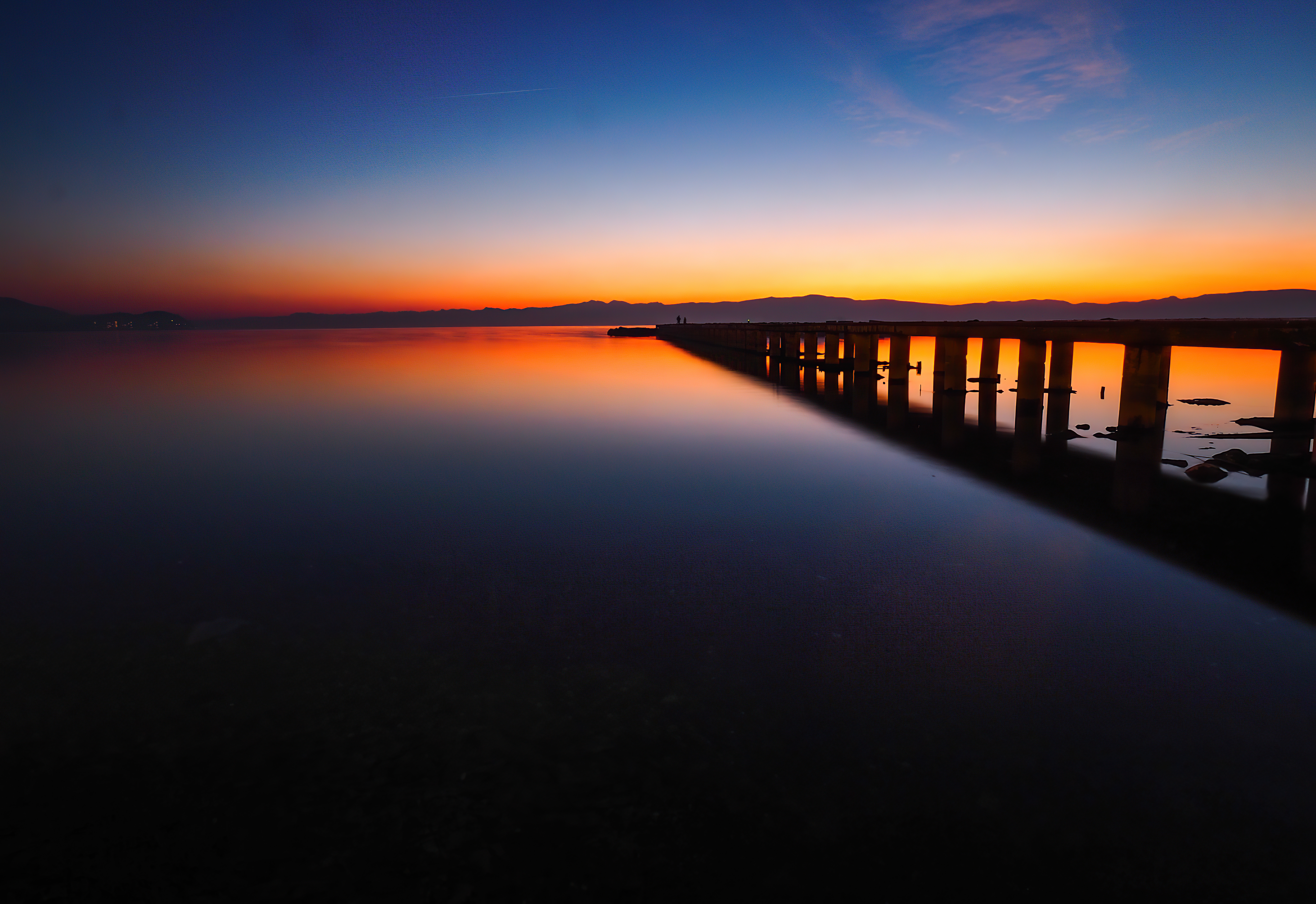Pier over calm water at sunset with mountains HD Wallpaper