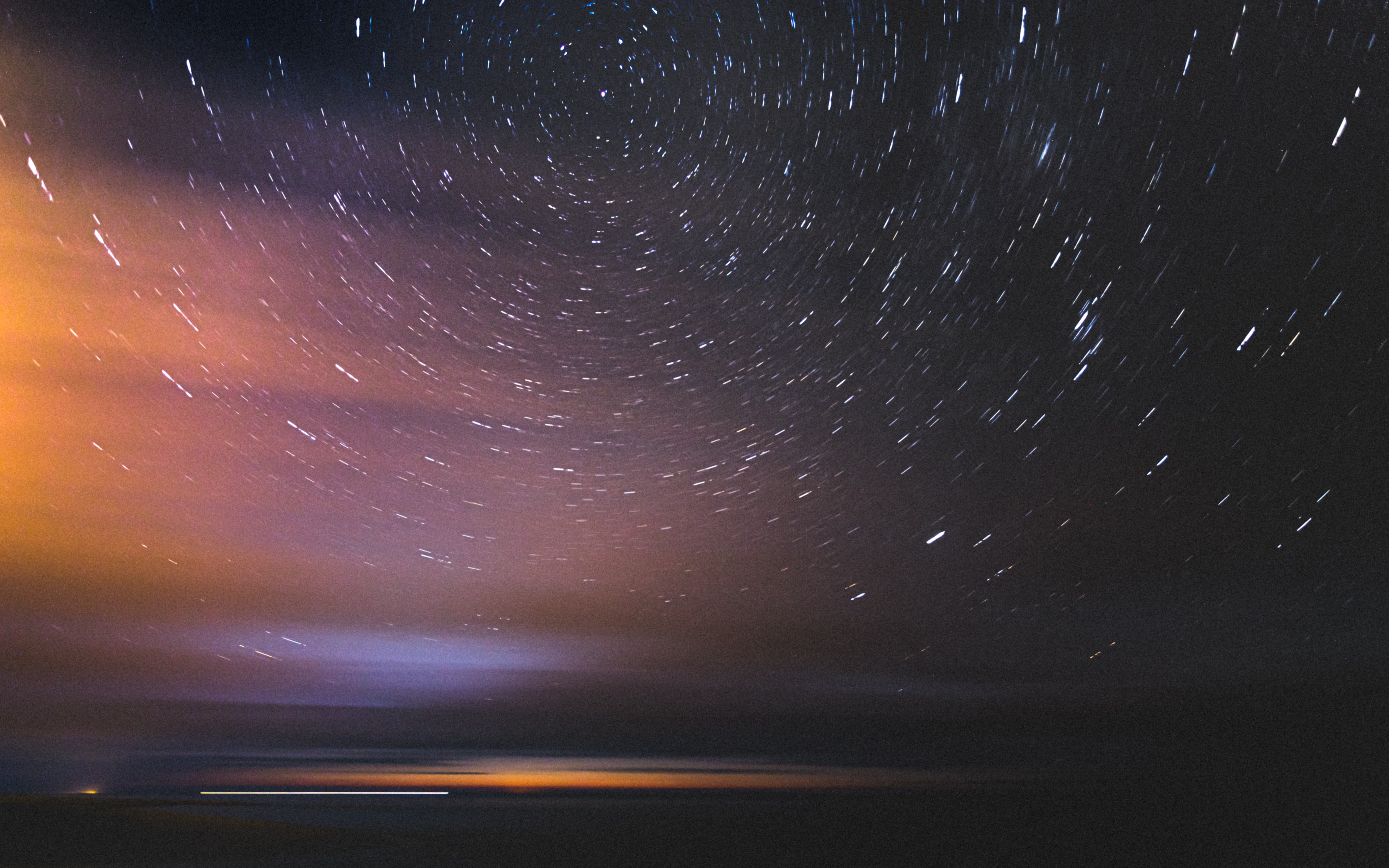 Star trails over a dark landscape with orange and purple clouds HD Wallpaper