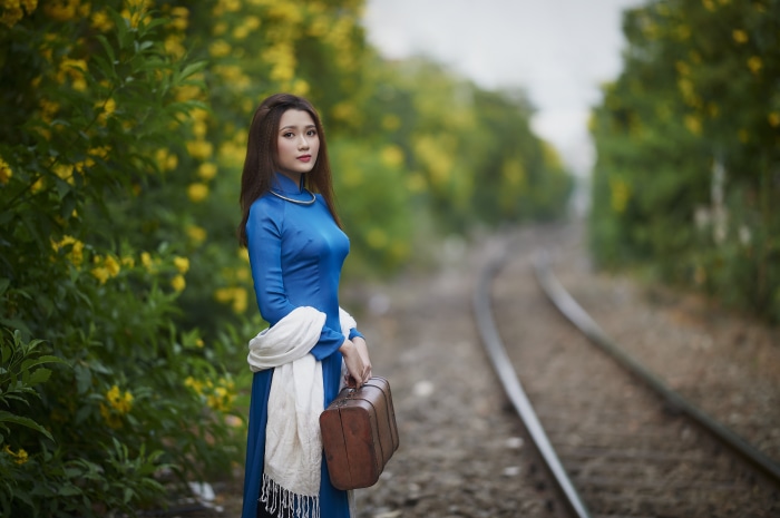Asian woman in blue dress with suitcase by train tracks