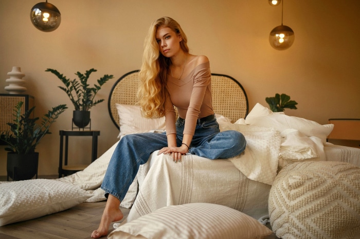 Woman in jeans and off-shoulder top sitting on a bed
