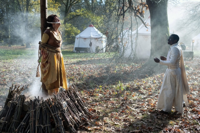 Woman tied to tree with fire pit, man in white robes