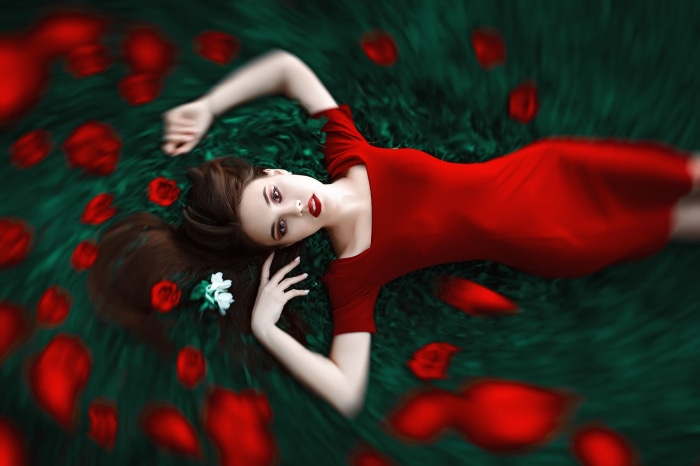 Brunette woman in red dress lying on grass with rose petals