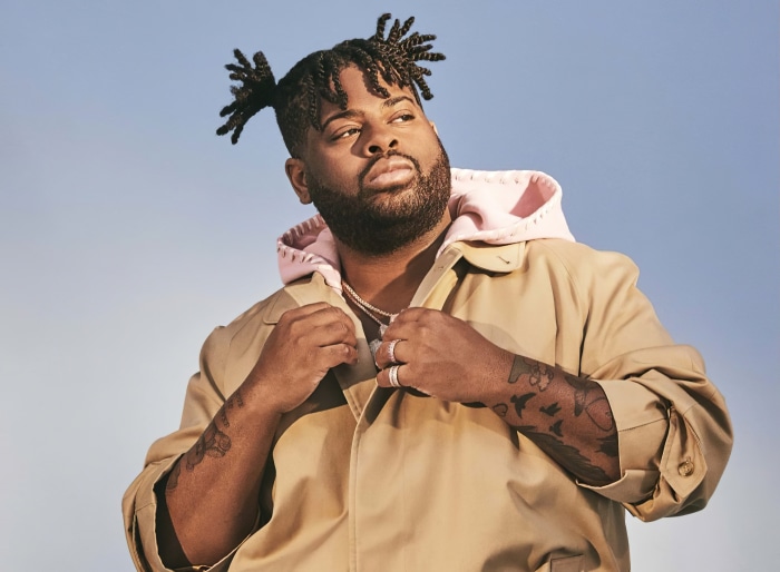 Man with dreadlocks wearing a tan jacket and pink hoodie