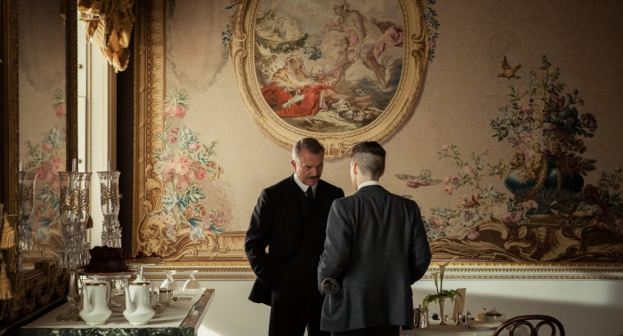 Two men in suits in a room with ornate wallpaper