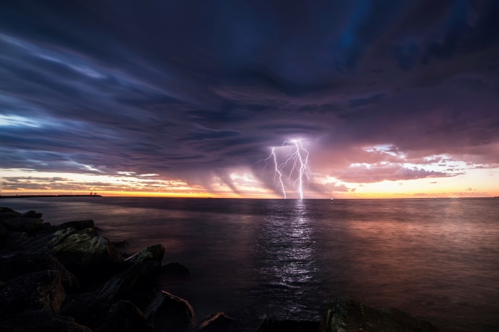Lightning strikes the ocean at sunset with rocky shore