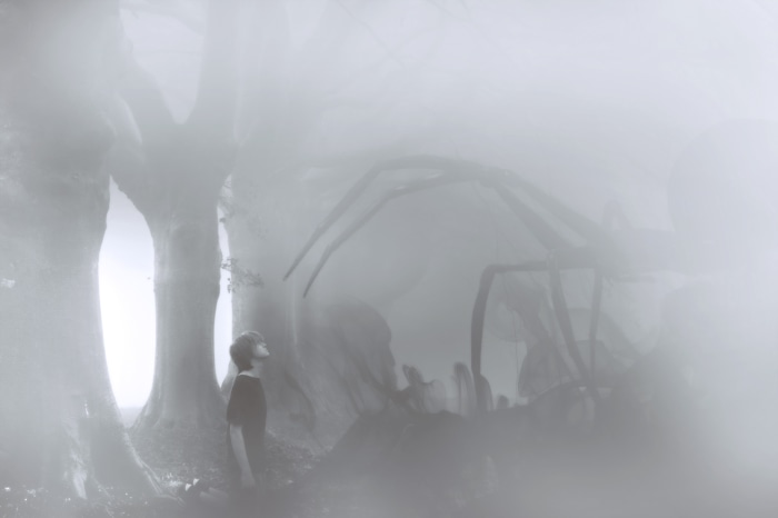 A child looks up at a giant spider in a foggy forest