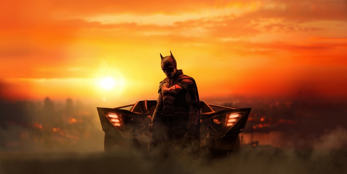 Batman from The Batman in front of the Batmobile at sunset