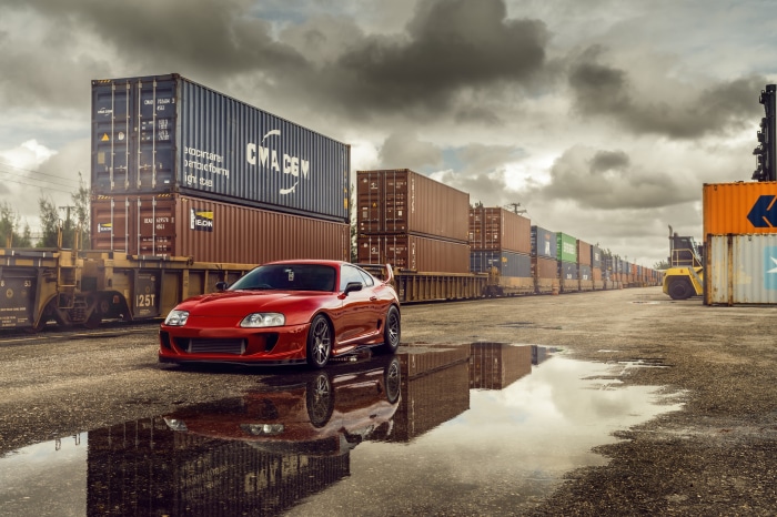 Red Toyota Supra parked by shipping containers