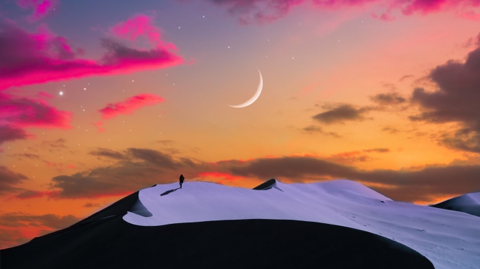 Person on sand dune under crescent moon and colorful sky