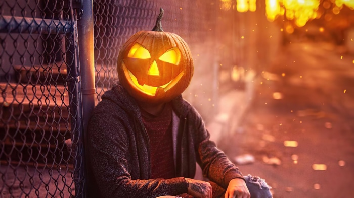 Person with Jack-o'-lantern head sitting by fence