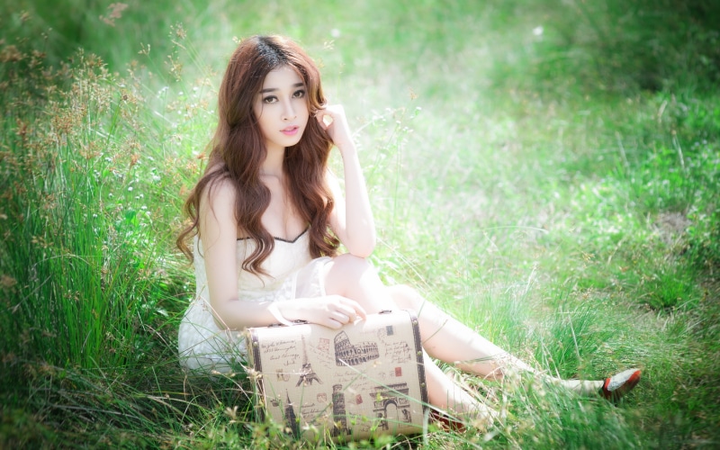 Asian woman with brown hair sitting in grass with suitcase