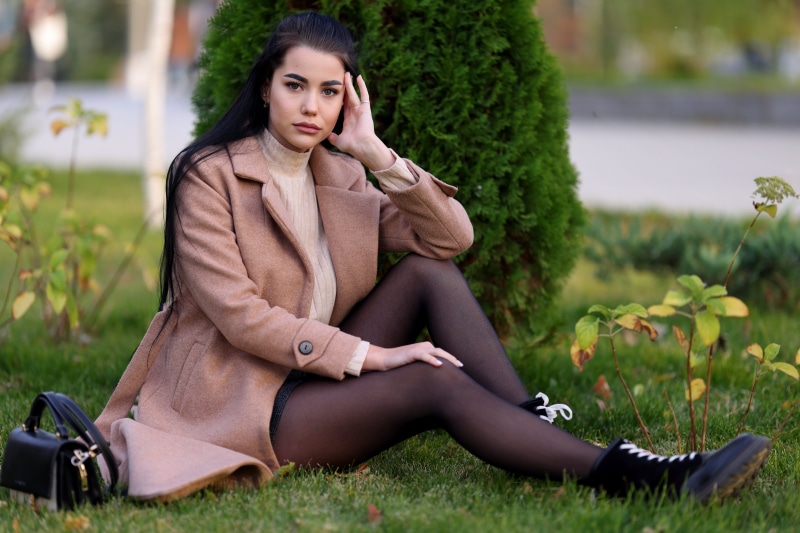 Brunette girl in coat sitting on grass with handbag