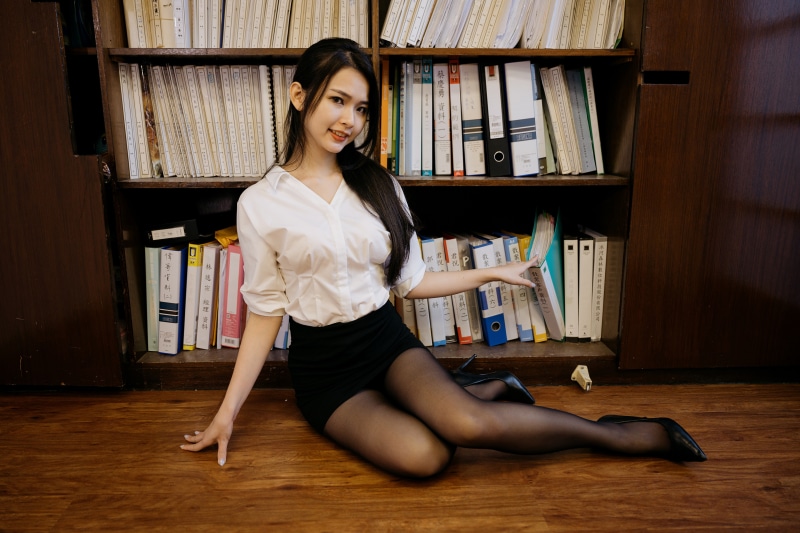 Asian woman in white blouse and black skirt sitting by bookshelf