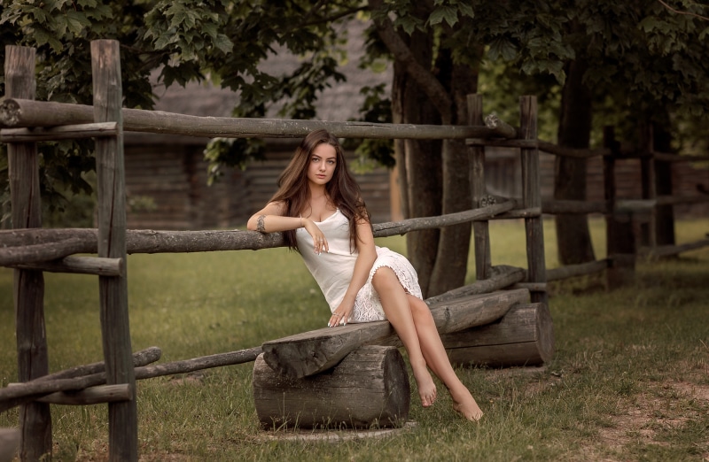 Woman in white dress sitting on a wooden bench near a fence