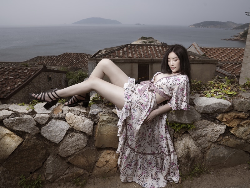 Asian woman in floral dress posing on stone wall overlooking village and sea