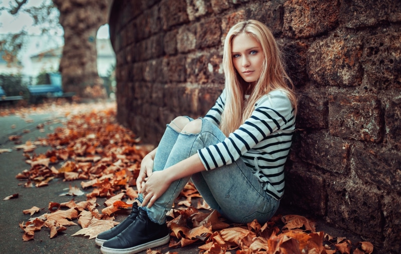 Blonde girl sitting by a brick wall with fallen leaves
