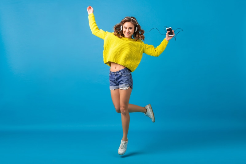 Woman in yellow sweater and denim shorts jumping with headphones and smartphone