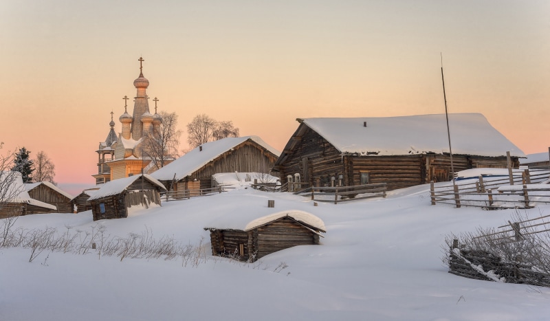 Wooden church and houses in a snowy Russian village at sunset