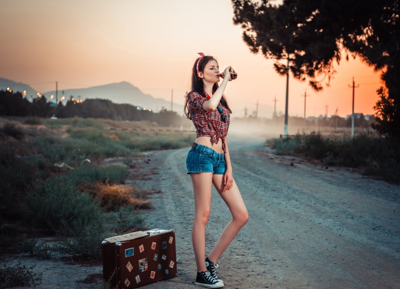 Woman drinking from bottle on rural road with suitcase