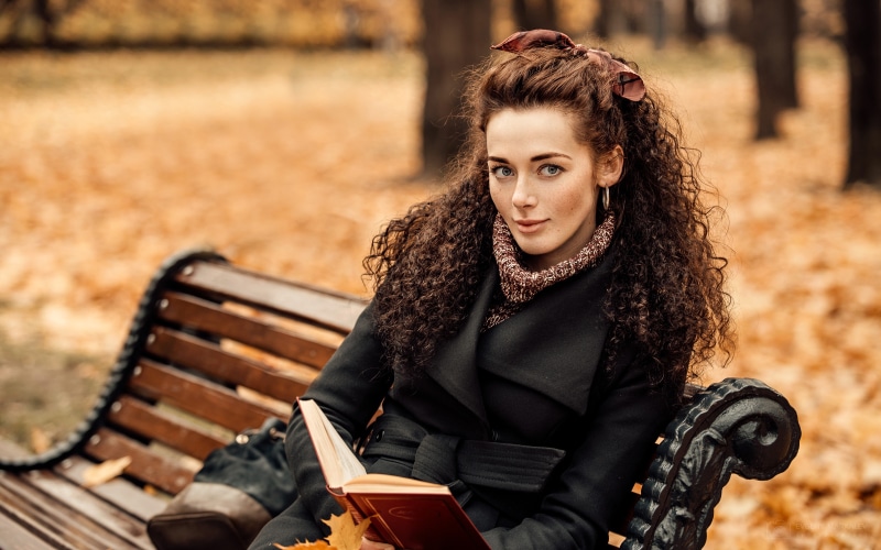 Woman with curly hair reading a book on a bench in autumn