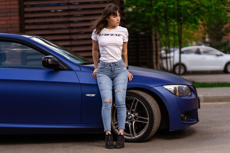 Woman in T-shirt and Jeans leaning on a blue car