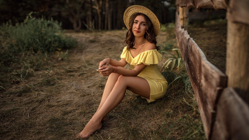 Woman in yellow dress and straw hat sitting by a fence