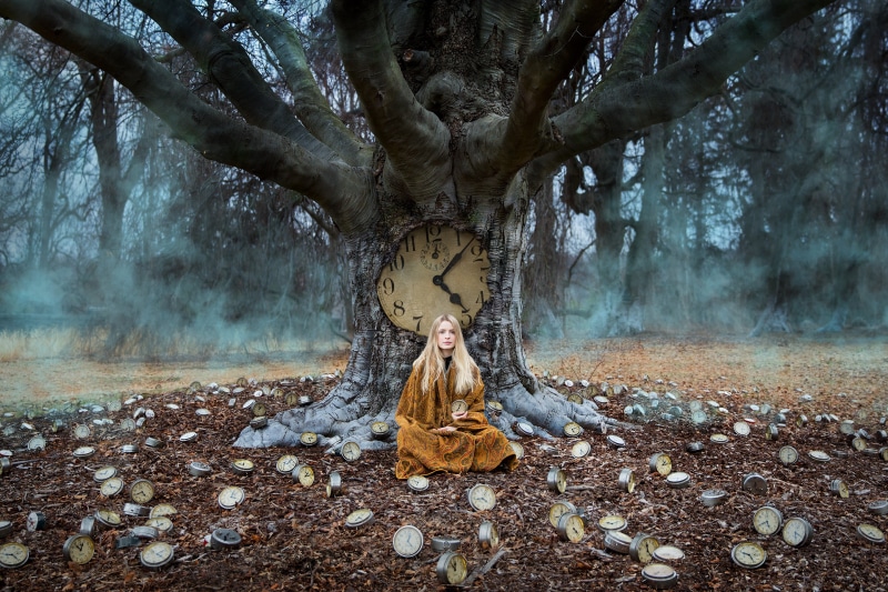 Blonde girl sitting by a tree with a clock and many clocks on the ground