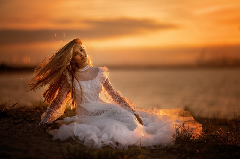 Blonde girl in white dress sitting by the water at sunset