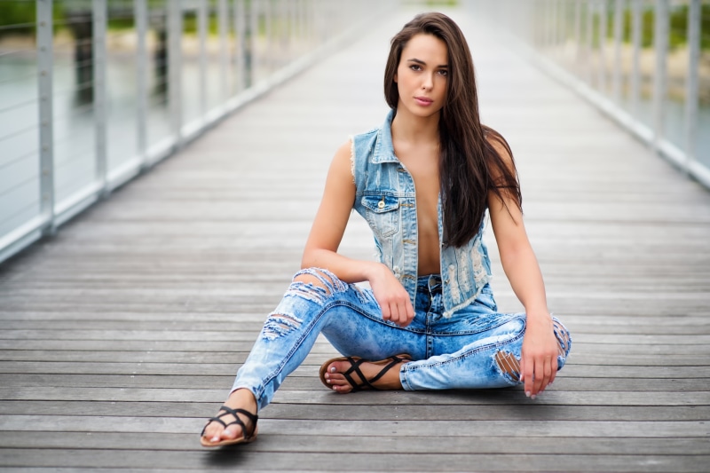 Woman in ripped jeans and denim vest sitting on boardwalk