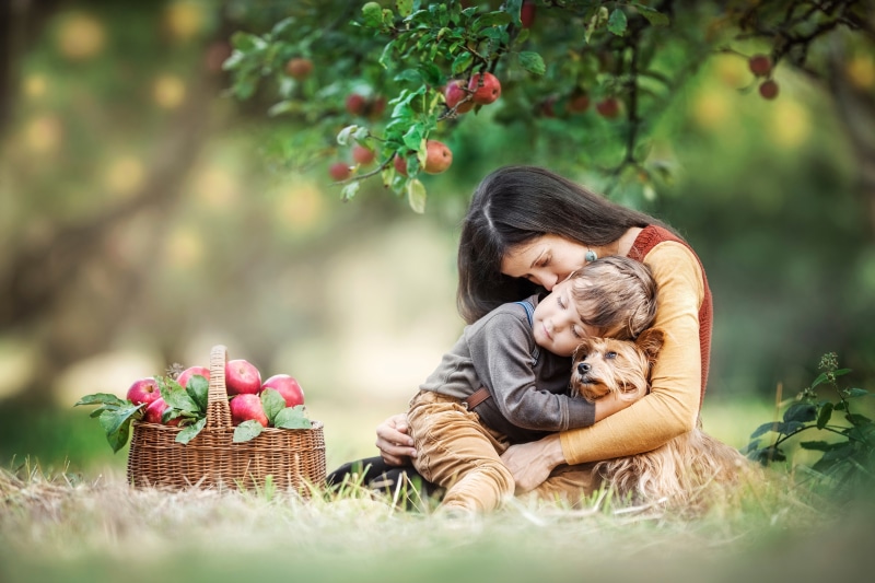 Mother hugging son and dog under apple tree