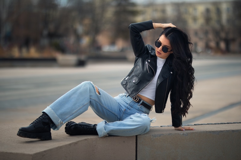 Brunette girl in leather jacket and ripped jeans posing outdoors