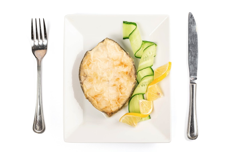 Fish steak with cucumber and lemon on a plate with fork and knife