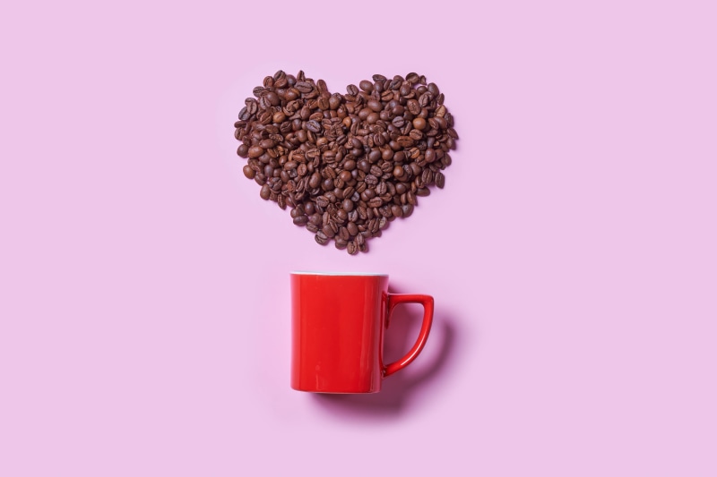 Coffee beans in heart shape above red mug on pink background