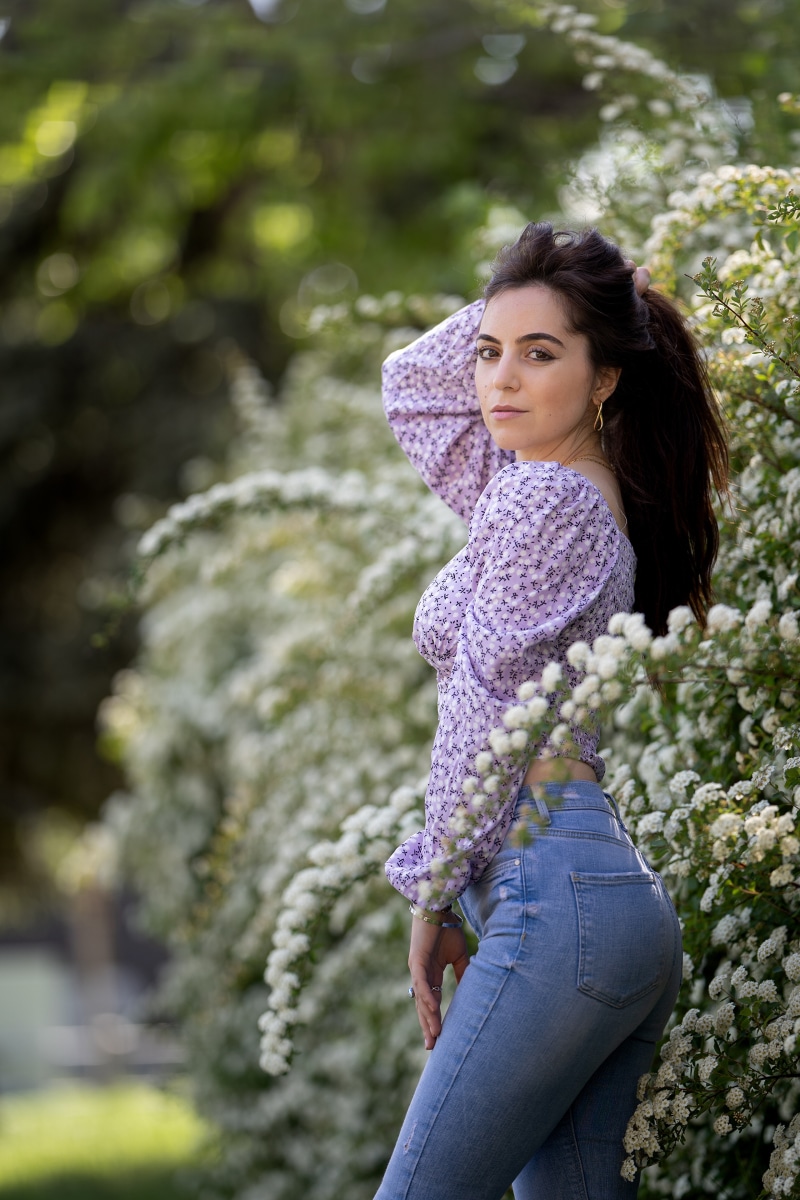 Brunette girl in jeans and floral top posing near white shrubs