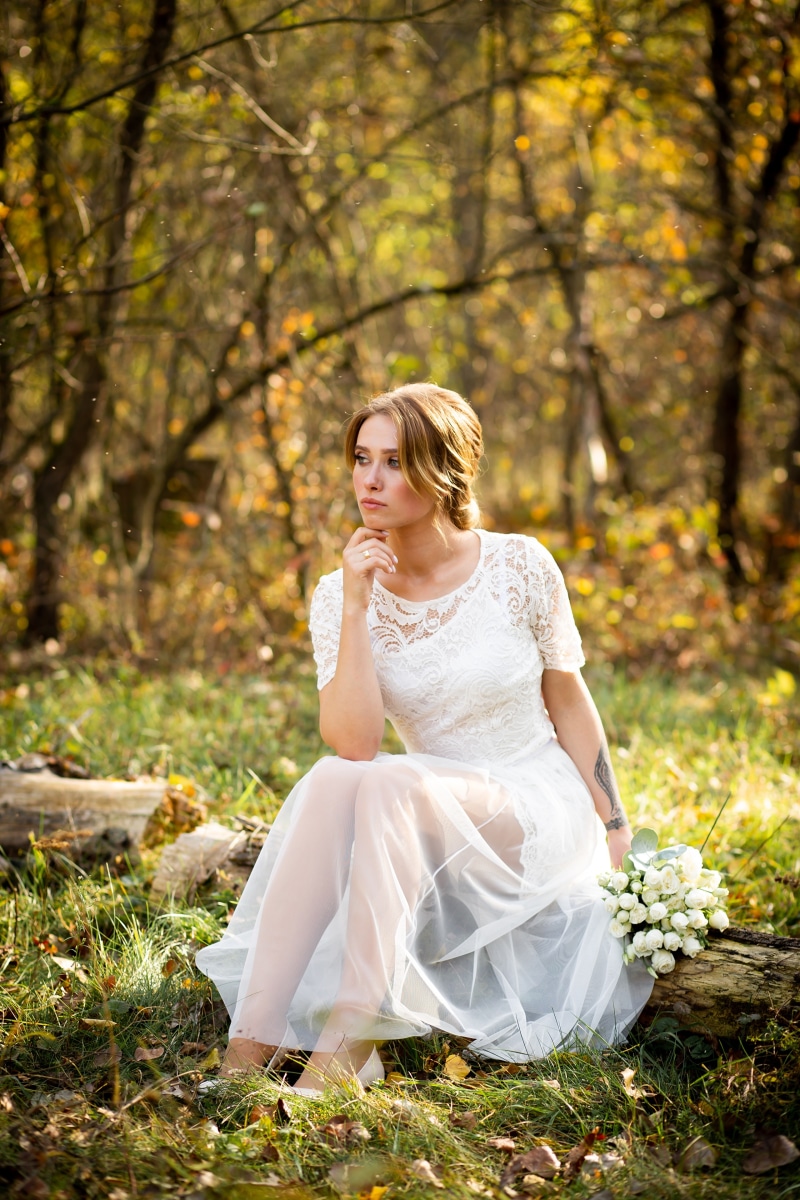Bride in white dress sitting in autumn forest with bouquet