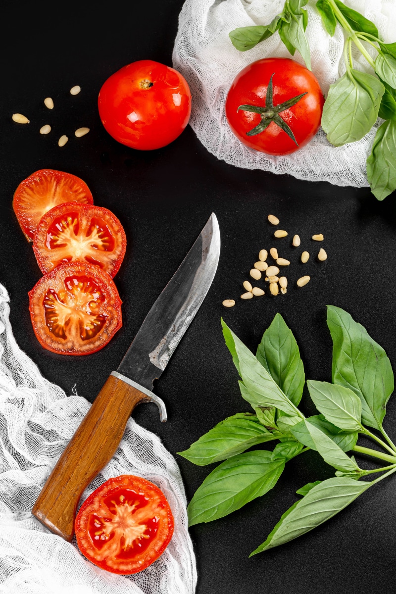 Tomatoes, basil, pine nuts, and a knife on a dark background